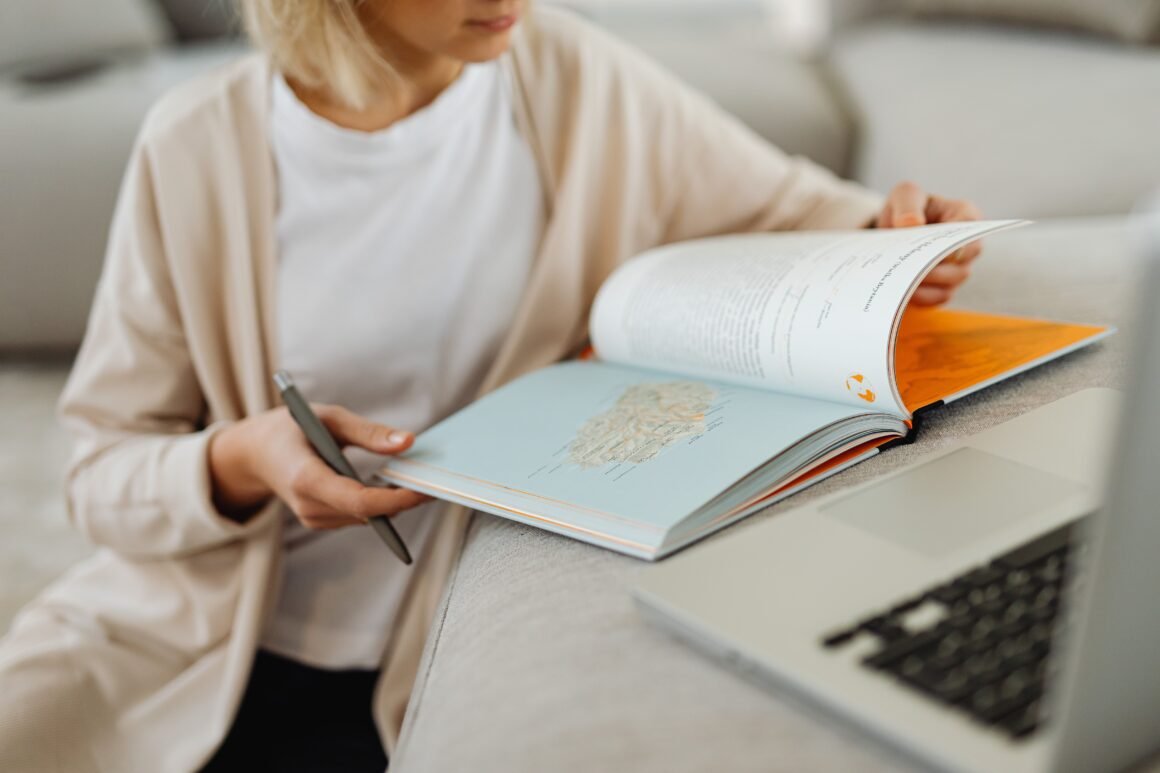 A person reading an open book beside a laptop on a sofa.