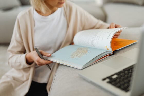 A person reading an open book beside a laptop on a sofa.