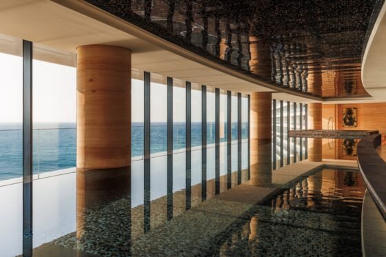 Sunlit indoor pool with warm stone columns, reflective ceiling and full-height windows overlooking the sea.
