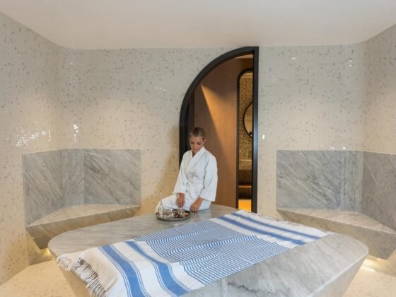 Pale mosaic and marble hammam with a central stone platform, striped cloth and a robed model beside an arched doorway.
