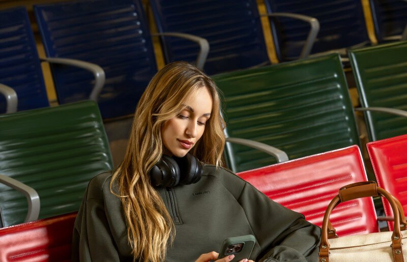 A woman in an olive travel top with headphones around her neck, seated among airport chairs.